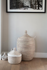Woven baskets with lids on a wooden floor against a light gray wall with a framed black and white photograph.