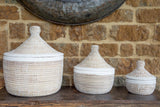 Three woven baskets with lids on a wooden surface against a brick wall.