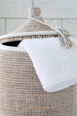 Woven laundry basket with a white towel draped over it against a light gray tiled wall.