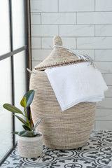 Handwoven white laundry basket with lid and tassel, styled with folded white towels.