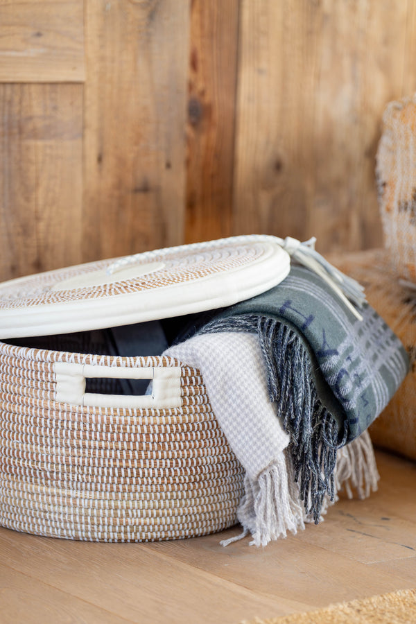 Woven storage basket with a lid and a blanket on a wooden floor.