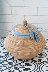 Woven basket with a blue lid and tassels on a tiled floor