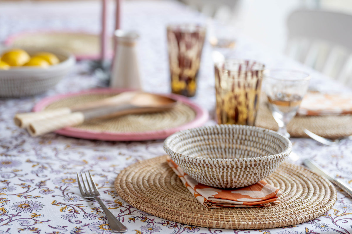 Dining table setting with woven bowl, plates, and cutlery on a floral tablecloth.