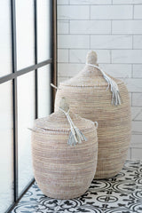 Two white handwoven laundry baskets with lids in medium and small sizes, shown on a tiled floor.