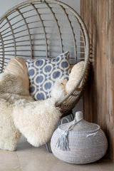 Wicker chair with fluffy cushions and a patterned pillow, next to a woven basket on a wooden floor.