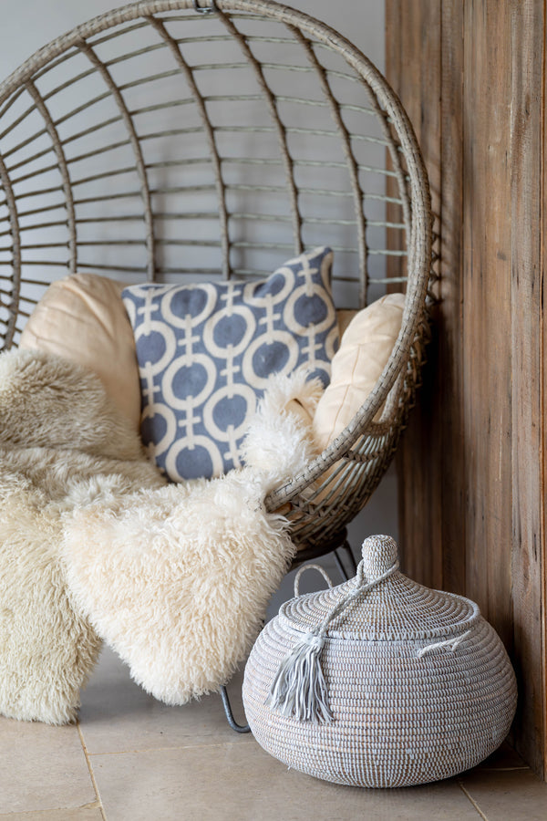 Wicker chair with fluffy cushions and a patterned pillow, next to a woven basket on a wooden floor.