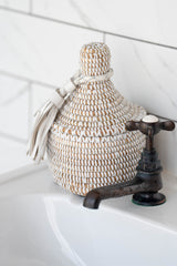 Small white handwoven storage basket with lid and leather tassel on bathroom sink
